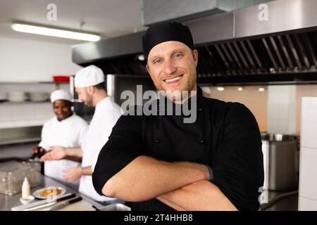 Vielseitig lächelnder männlicher Chefkoch mit überkreuzten Armen, der vor Studenten steht, die im Hintergrund kochen Stockfoto