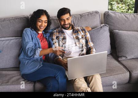 Glückliches birassisches Paar, das auf dem Sofa sitzt und im Wohnzimmer zu Hause einen Laptop benutzt Stockfoto