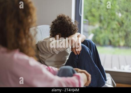 Ein verärgerter Junge sitzt auf dem Sofa, während seine Mutter ihn ansieht Stockfoto