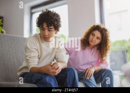 Ein verärgerter Junge sitzt auf dem Sofa, während seine Mutter ihn ansieht Stockfoto