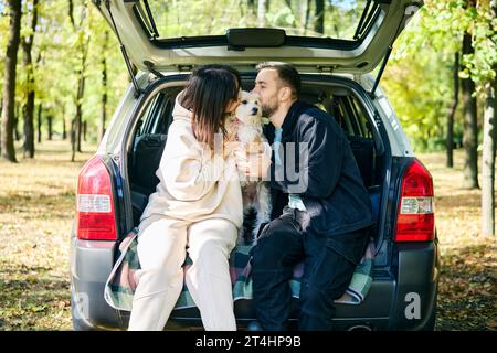 Ein Paar küsst liebevoll ihren liebenswerten Hund, der im Kofferraum sitzt und sich im Wald entspannt. Familie, Liebe, Urlaub und Reisekonzept Stockfoto