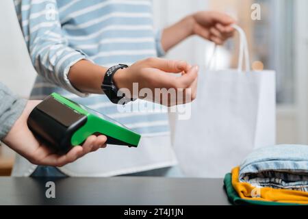 Moderne Frau, die ein Terminal für kontaktloses Bezahlen mit Smartwatch am Tresen im Bekleidungsgeschäft benutzt. NFC-Technologie. Weibliche Kundin, die den Kauf über Smart Watch bezahlt Stockfoto