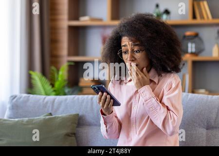 Traurig enttäuschte Frau erhielt am Telefon Nachrichten, afroamerikanerin las schlechte Nachrichten online mit App auf Smartphone, saß zu Hause auf Sofa im Wohnzimmer des Hauses. Stockfoto
