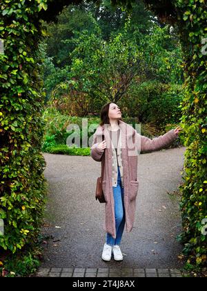 Junge Frau mit schulterlangen braunen Haaren, die an einem Herbsttag in Museum Gardens in York Yorkshire, England, unter einem Buchenbogen stand Stockfoto