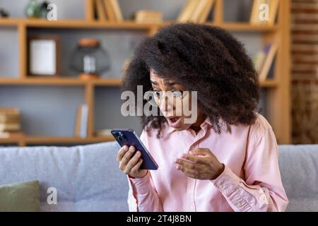 Traurig enttäuschte Frau erhielt am Telefon Nachrichten, afroamerikanerin las schlechte Nachrichten online mit App auf Smartphone, saß zu Hause auf Sofa im Wohnzimmer des Hauses. Stockfoto