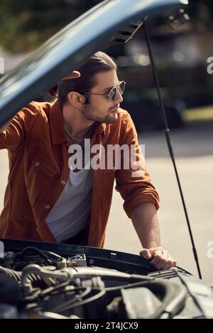 Gut aussehendes männliches Modell mit Sonnenbrille, die neben der geöffneten Motorhaube steht und wegblickt Stockfoto