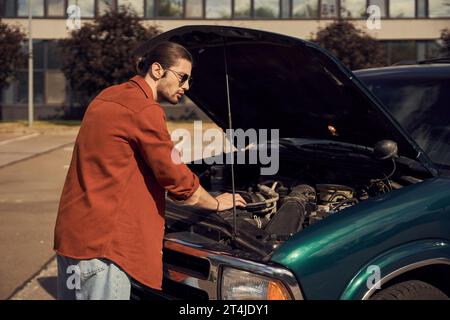 Schöner bärtiger Mann mit Pferdeschwanz und Sonnenbrille neben seinem Auto mit geöffneter Motorhaube Stockfoto