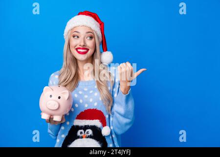 Foto-Porträt der attraktiven blonden Frau Punkt leerer Raum Schweinchen gekleidet Pinguin Pullover x-MAS Hut isoliert auf blauem Farbhintergrund Stockfoto