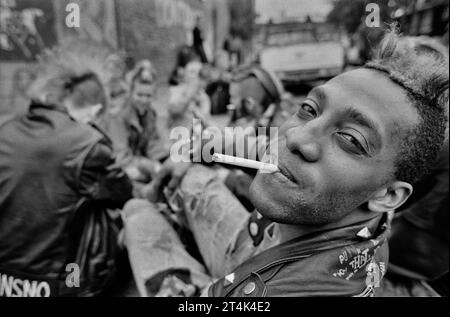 Ein junger schwarzer Punk mit einer Gruppe von Freunden, die auf einem Brixton-Pflaster sitzen. Stockfoto