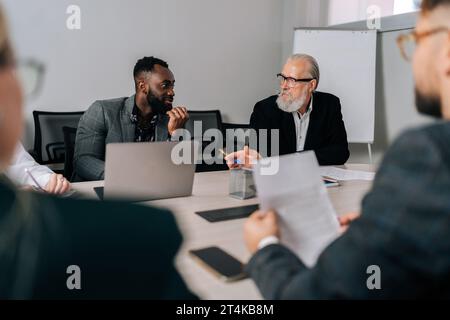 Blick von der Schulter auf den afrikanischen Teamleiter, der am Tisch sitzt und beim Briefing-Meeting Projektideen mit Investoren aus gemischten Rassen teilt. Stockfoto