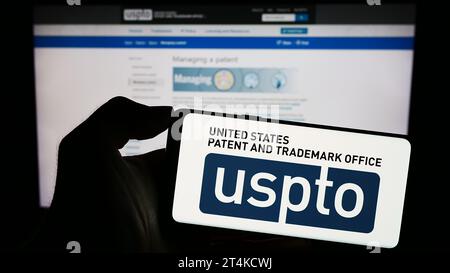 Person, die ein Mobiltelefon mit dem Logo der Agentur United States Patent and Trademark Office (USPTO) vor der Webseite hält. Schwerpunkt auf dem Display des Telefons. Stockfoto