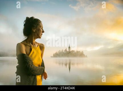Frau in gelbem Kleid, die einen Sonnenaufgang im Nebel am Ufer des Bleder Sees in Slowenien beobachtet Stockfoto