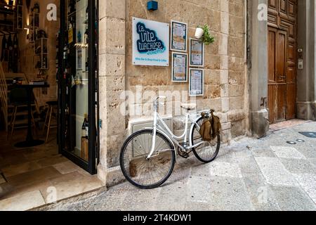 Bezaubernde Weinbar in Monopoli, Italien, mit einem alten weißen Fahrrad und Menüs auf einer rustikalen Steinmauer entlang einer engen Kopfsteinpflasterstraße. Stockfoto