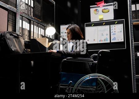 Asiatischer Wirtschaftsprüfer im Rollstuhl, der administrative analytische Daten im Kabinettsbüro überprüft, umgeben von Buchführungsstatistiken. Geschäftsfrau mit Behinderung im Aktenlager Stockfoto