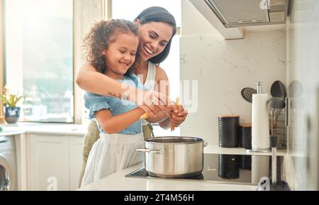 Mutter und kleine Tochter kochen gemeinsam in der Küche. Mutter und Kind aus gemischter Rasse stehen am Herd, brechen Spaghetti und werfen sie hinein Stockfoto