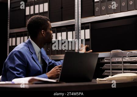 afroamerikanischer Offizier, der Regale mit Polizeidokumenten ansieht, auf der Suche nach Expertise der Kriminologie-Abteilung im Arhive-Raum. Privatdetektive, die Überstunden im Strafverfahren arbeiten Stockfoto