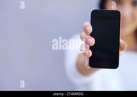 Du musst dieses Telefon holen. Porträt einer jungen Frau, die vor grauem Hintergrund steht und ihr Handy hochhält. Stockfoto