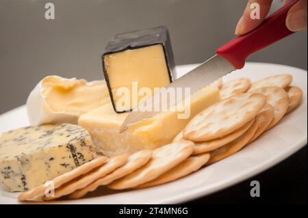 Eine Auswahl an Käsesorten, auf einem Teller mit einfachen Crackern und einer Hand mit Messer. Stockfoto