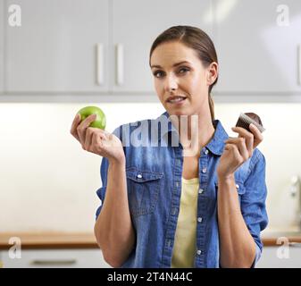 Die richtige Wahl zu treffen, ist nicht einfach... Eine Frau, die versucht, zwischen einem Cupcake und einem Apfel zu wählen. Stockfoto