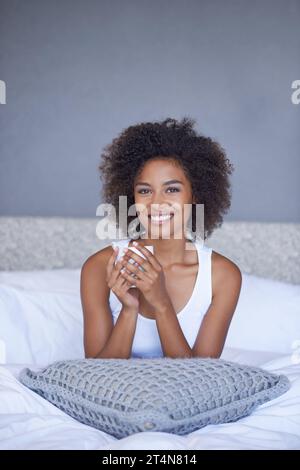 Es ist nichts Schöneres, als mit Kaffee in der Tasse aufzuwachen. Eine junge Frau genießt eine Tasse Kaffee, während sie auf dem Bett liegt. Stockfoto