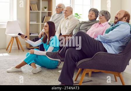 Glückliche Seniorinnen sehen Fernsehen, sitzen auf dem Sofa im Altersheim mit einer jungen Krankenschwester. Stockfoto