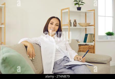 Porträt einer glücklichen, selbstbewussten Frau, die zu Hause auf der Couch sitzt, in die Kamera blickt und lächelt Stockfoto
