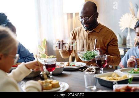 Glücklicher junger Mann, der selbstgemachten Gemüsesalat auf seinen Teller legt, während er mit seiner Frau und seinen Töchtern beim festlichen Abendessen im Wohnzimmer sitzt Stockfoto
