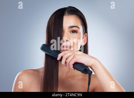 Schöne junge Frau lächelt, während sie einen Haarglätter auf ihrem langen braunen Haar benutzt. Brünette Frau mit Flacheisen. Verwenden von Haarpflegeprodukten zu Stockfoto