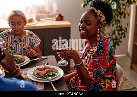 Glückliche junge afroamerikanische Frau in ethnischer Kleidung, die während des Nationalfeiertags Weinglas mit rotem hausgemachtem Wein hält Stockfoto