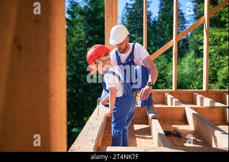 Vater mit Kleinkind-Sohn, der ein Holzrahmenhaus baut. Vater Bauarbeiter, der seinem Sohn an sonnigen Tagen die Fähigkeit vermittelt, Nägel mit Hammer zu fahren. Zimmerei und Familienkonzept. Stockfoto