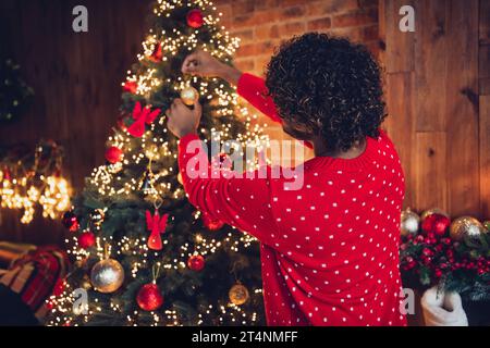 Rückansicht des funkigen jungen, nicht erkennbaren arabischen Mannes, der roten Pullover trägt und Fichtenbaum als traditionelle Party für ein glückliches neues Jahr dekoriert Stockfoto
