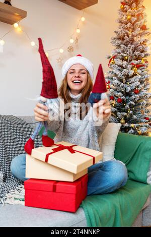 Junge Frau mit Weihnachtsmütze, die zu Hause auf der Couch sitzt und Weihnachtsgeschenke auspackt. Stockfoto
