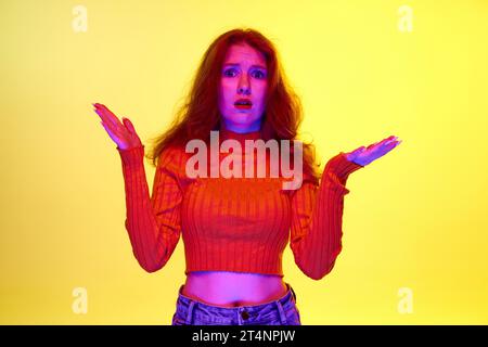 Junges Mädchen, das mit unglücklichem Gesicht steht und Hände mit Schock vor gelbem Atelierhintergrund im Neonlicht spreizt Stockfoto