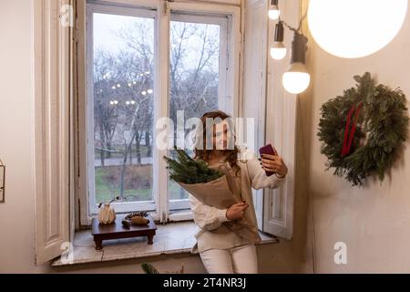 Stilvolle Frau in weißem Pullover nimmt Selfie auf dem Smartphone auf, hält Weihnachtsstrauß in der Hand in Bastelpapier in der Nähe des Fensters. Junge Millennials Mädchen mit f Stockfoto