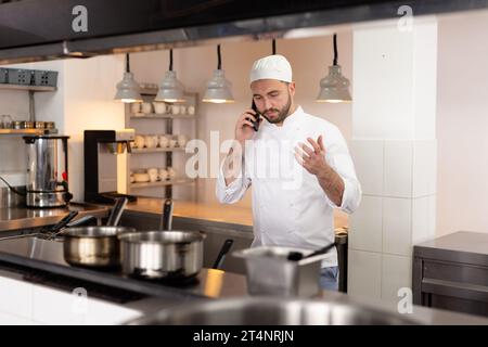 Konzentrierter kaukasischer Koch, der über Smartphone spricht, gestikuliert in der Küche des Restaurants, Kopierraum Stockfoto