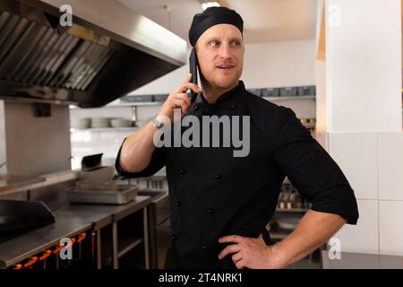 Glücklicher kaukasischer Küchenchef, der auf dem Smartphone mit Hand auf Hüfte in der Restaurantküche spricht, Kopierraum Stockfoto