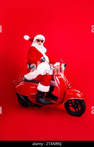 Foto in voller Größe des erstaunten lustigen Großvaters tragen trendige santa Kostümbrille, die schnell auf Moped fährt, isoliert auf rotem Hintergrund Stockfoto