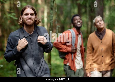 Porträt eines jungen Mannes mit Rucksack, der in die Kamera schaut, während er zusammen mit seinen Freunden im Wald spaziert Stockfoto