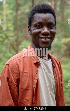 Vertikales Porträt eines afroamerikanischen Mannes, der in die Kamera lächelt, während er im Wald spaziert Stockfoto