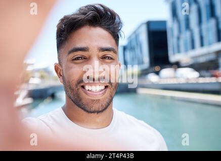 Junger Mixed-Race-Sportler lächelt und hält Handy mit der Hand, um ein Selfie zu machen, während er auf einem Wasserkanal in der Stadt läuft oder joggt Stockfoto