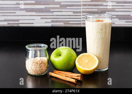 Ein Glas mit hausgemachtem Smoothie neben einer halben Zitrone, einem grünen Apfel, einem Glastopf mit Haferflocken und zwei Zimtstangen über einer schwarzen Küchenbank. Stockfoto