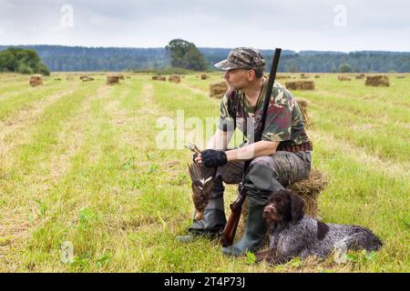 Ein Jäger sitzt auf einem gemähten Heu und hält ein abgefallenes Auerhühnchen in den Händen, ein Jagdhund liegt neben ihm Stockfoto