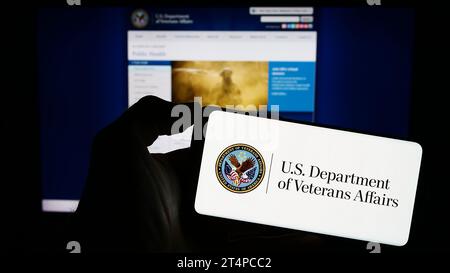 Person, die vor der Webseite ein Mobiltelefon mit dem Siegel des United States Department of Veterans Affairs (VA) hält. Schwerpunkt auf dem Display des Telefons. Stockfoto