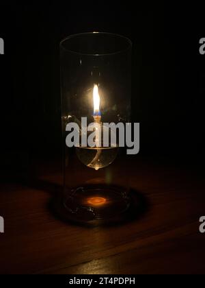 Leuchtende Öllampe aus klarem Glas, die in dunkler Umgebung auf einem Tisch platziert wird und ein Leuchten mit dunklem Hintergrund erzeugt. Stockfoto