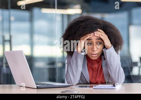 Frustrierte und müde depressive Geschäftsfrau, die am Arbeitsplatz im Büro sitzt, unglückliche afroamerikanische Frau im Business-Anzug, die mit Laptop arbeitet, gescheitert und bankrott. Stockfoto