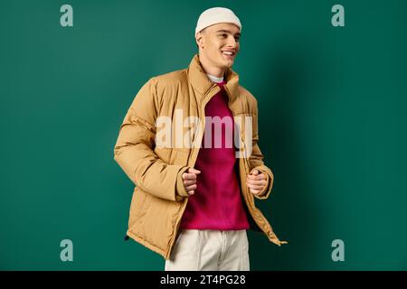 Fröhlicher junger Mann in Mütze und stilvoller Pufferjacke auf türkisfarbenem Hintergrund, Wintertrends Stockfoto