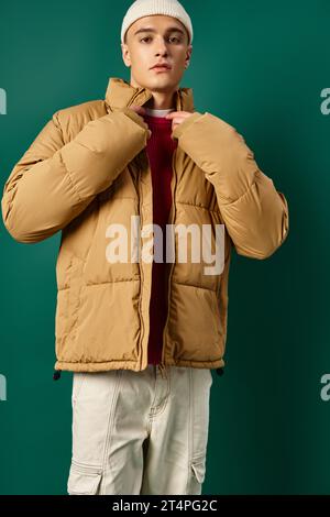 Junger Mann in Beanie-Hut mit stilvoller Pufferjacke auf türkisfarbenem Hintergrund, Wintertrends Stockfoto