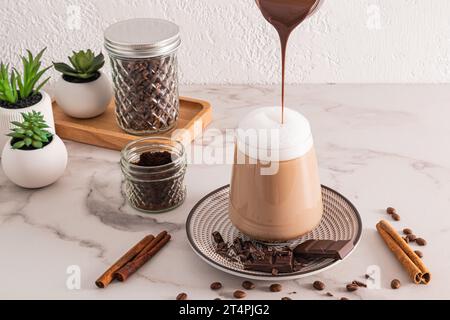 Ein Hauch heißer Schokolade wird in ein großes Glas Kaffee, Cappuccino oder Zimt Latte gegossen. Vorderansicht mit Marmorhintergrund. Ein schickes Dessert Stockfoto