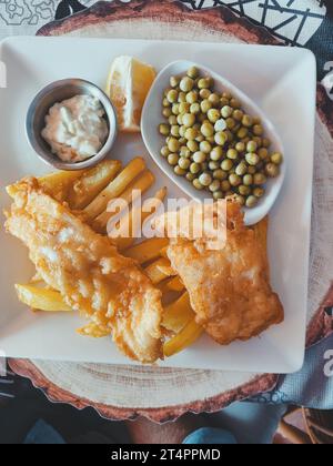 Vertikales Foto von Draufsicht von Fisch mit Pommes Frites und grünen Erbsen auf einer weißen quadratischen Platte mit Saucen. Weichzeichner. Kopierbereich Stockfoto