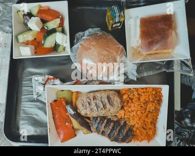 Draufsicht auf ein Tablett mit Lebensmitteln in einem Flugzeug. Zwei Schnitzel mit Bulgur, ein Brötchen, Olivenöl und frischem Gemüsesalat. Weichzeichner. Stockfoto